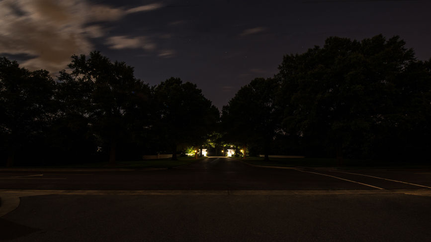 A dark, empty street at night with large trees on both sides and distant lights illuminating a house in the background.