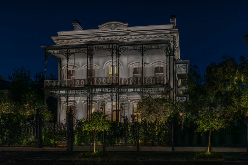 Two-story historic mansion with ornate iron balconies and columns, illuminated at night, surrounded by trees and a black wrought iron fence.