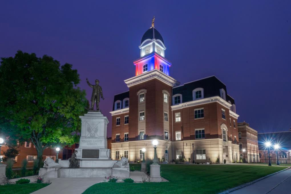 A brick courthouse with an illuminated dome in red, white, and blue lights at dusk; a statue stands on a pedestal in the landscaped lawn in the foreground.