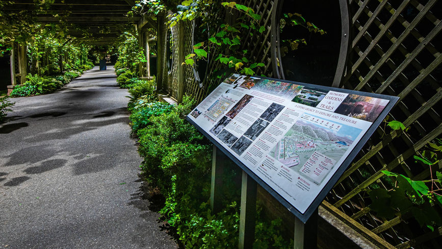 An informational sign with maps and text stands beside a shaded garden pathway lined with green plants and trellises.