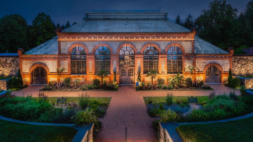 A large, historic glass and brick conservatory is lit from below at dusk, with arched windows, symmetrical gardens, and benches in the foreground.
