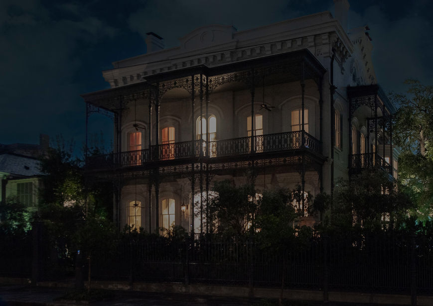 A large historic house with ironwork balconies and glowing windows is seen at night, partially obscured by trees and a fence in the foreground.