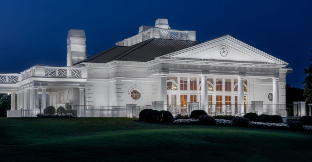 A large white neoclassical building with columns and a clock above the entrance is illuminated at night, standing on a manicured lawn.