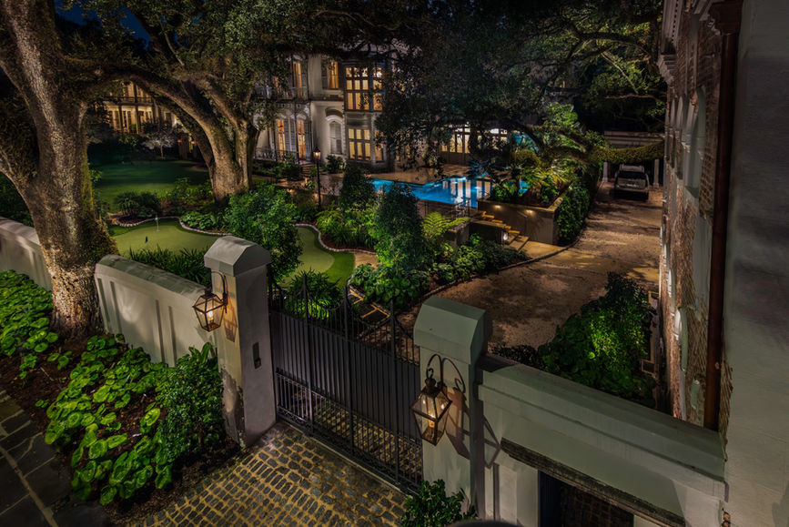Gated entrance to a large, well-lit house with manicured gardens, mature trees, and a backyard pool at night.