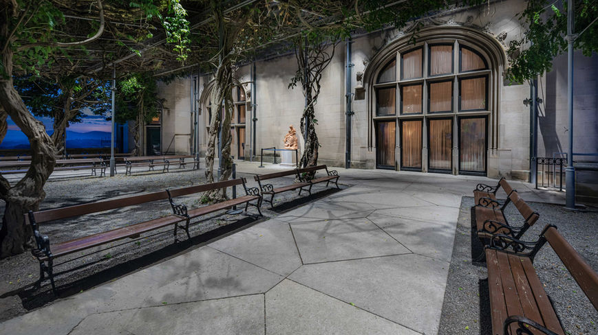 Outdoor courtyard with wooden benches, vine-covered pergola, large arched windows, and a statue in front of a stone building at dusk.