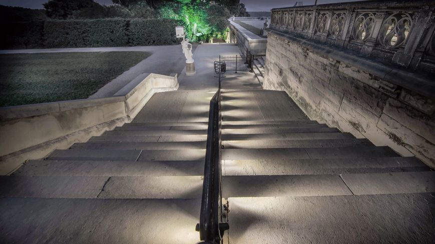 Stone steps with a metal handrail descend at night, illuminated by lights; a statue and green-lit bushes are visible in the background.