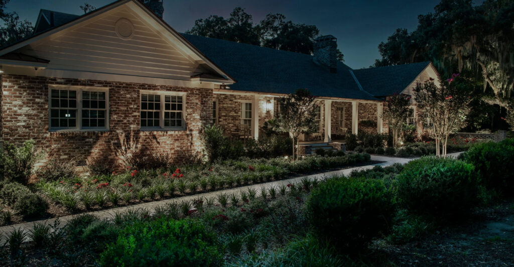 A brick house with a landscaped front yard and illuminated windows at night. Shrubs and small trees line the walkway leading to the front entrance.