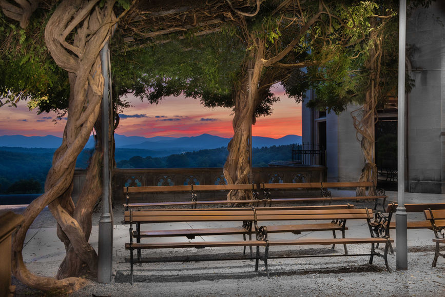 Wooden benches sit beneath twisted tree branches on a terrace, overlooking distant mountains at sunset with a colorful sky.