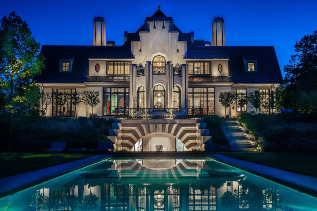 A large, illuminated two-story house with geometric architectural features is reflected in a rectangular swimming pool at night.