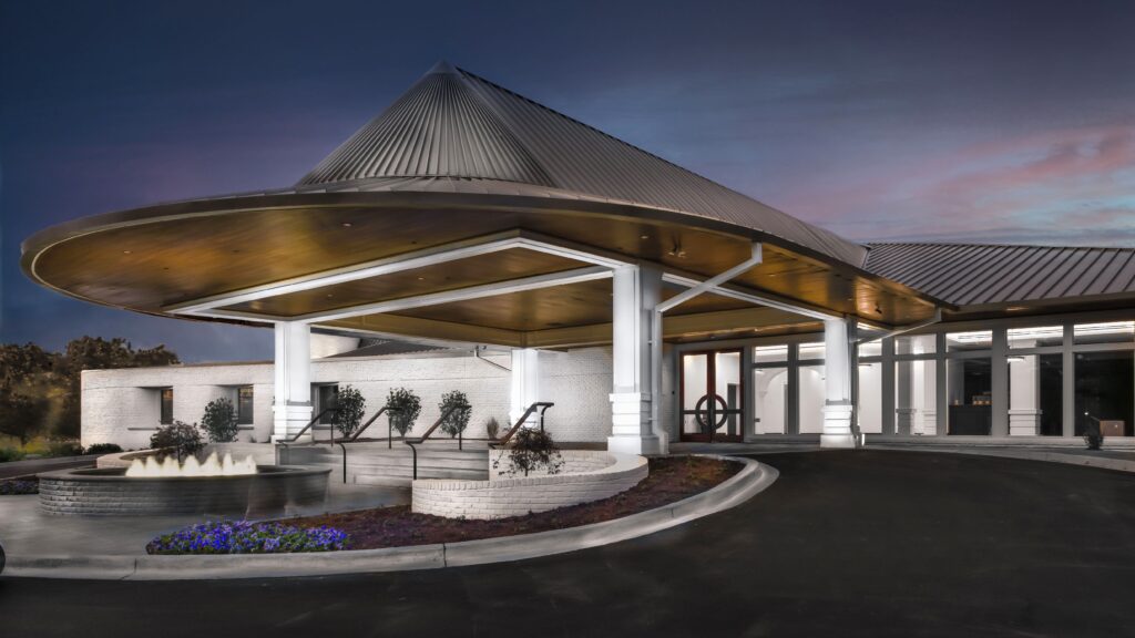 Modern building with a metal roof and covered entrance, featuring white columns, a small circular fountain, and landscaped flower beds in the foreground at dusk.