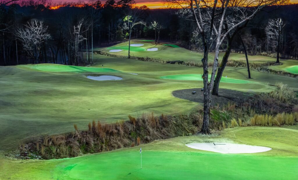 A golf course with sand bunkers and putting greens is shown at sunset, with trees and a dark forested background.